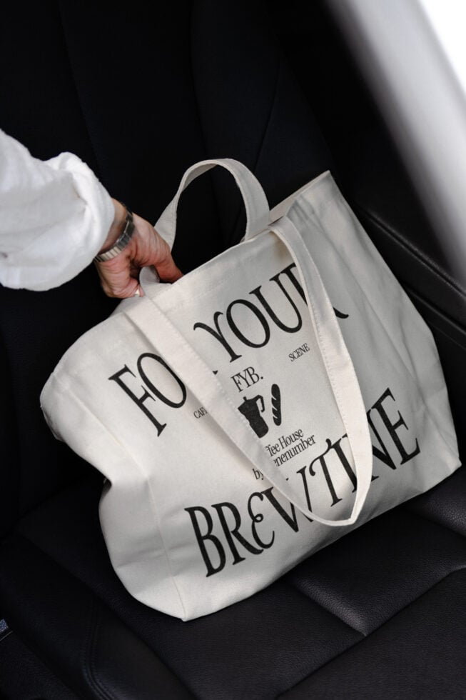 Close-up of white tote bag with For Your Brewtine typography being held against black background