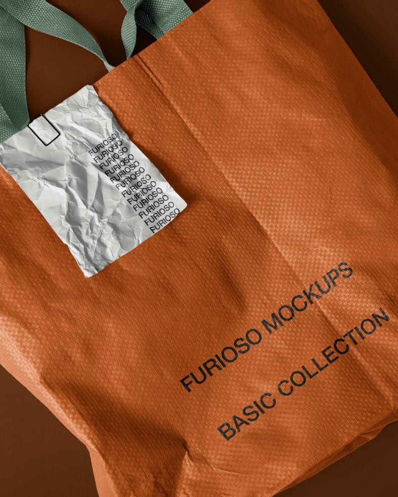 Orange fabric tote with crumpled white label tag, green handles, flat lay overhead shot