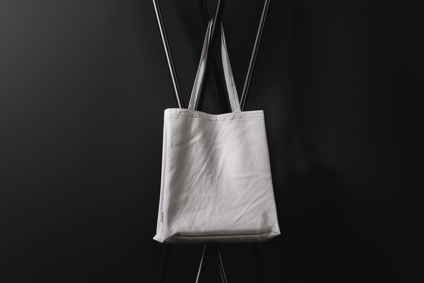 Blank white canvas tote bag hanging against black background with straps