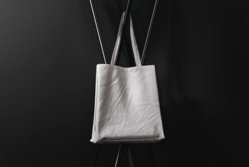 Blank white canvas tote bag hanging against black background with straps