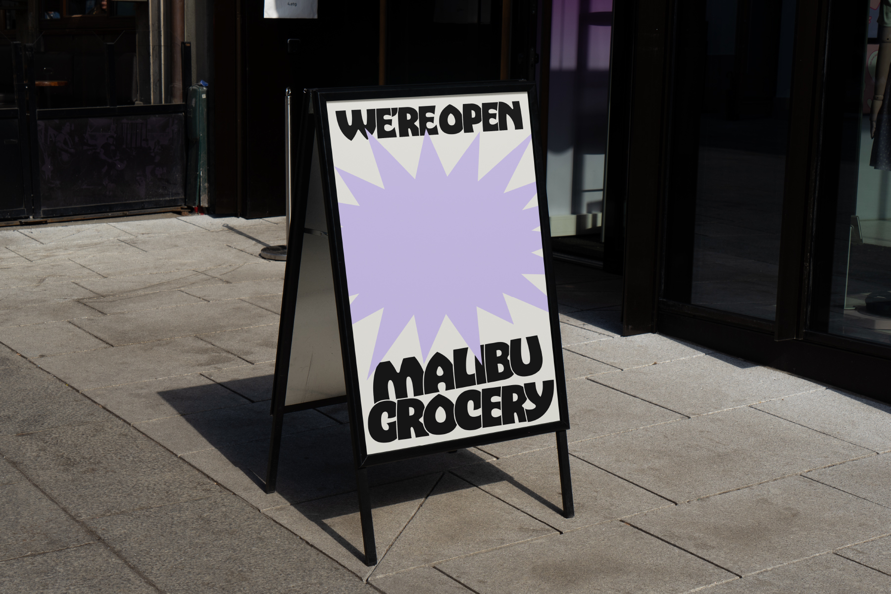 A-frame sandwich board on sidewalk showing we're open sign for malibu grocery with purple starburst