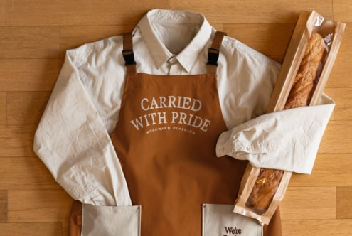 Brown apron with Carried With Pride text flat lay with bread, white shirts