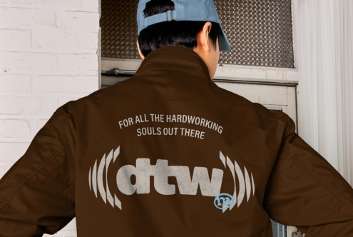 Brown jacket back with workwear logo, blue cap indoors by white brick