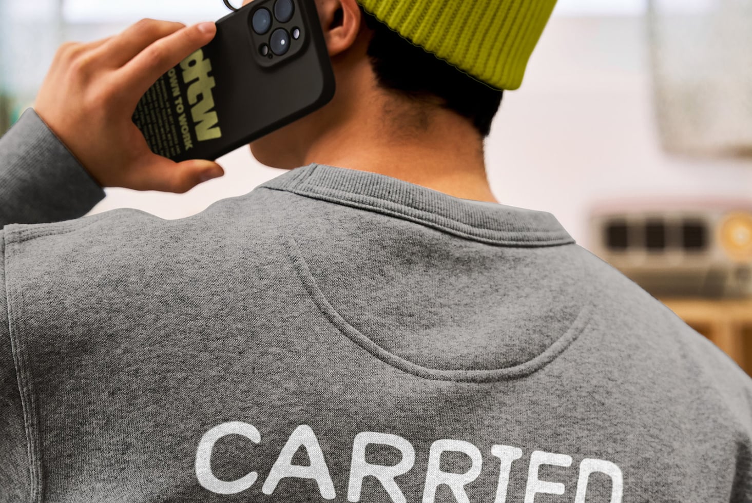 Back view of person in yellow beanie holding black phone with gray crewneck visible