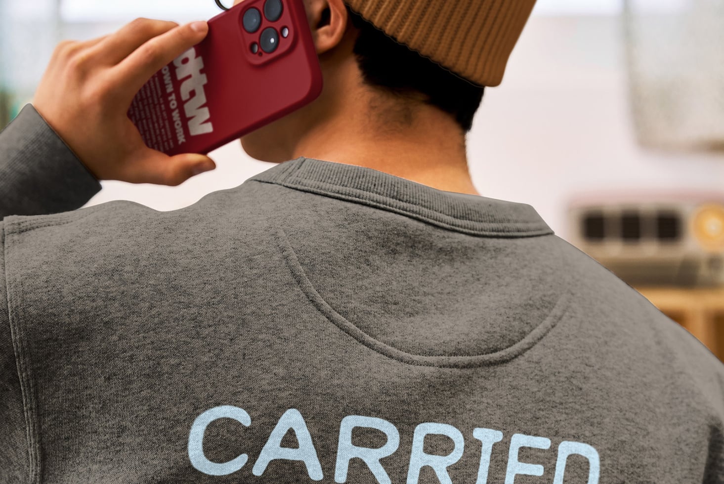 Person in brown beanie holding red phone case wearing gray crewneck with carried text