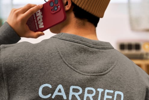 Person in brown beanie holding red phone case wearing gray crewneck with carried text