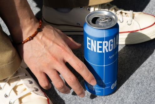 Hand with beaded bracelet holding blue Energia can on grey fabric with sneaker