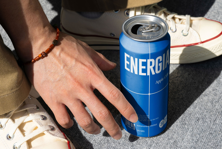 Hand with beaded bracelet holding blue Energia can on grey fabric with sneaker