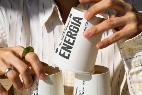 Hands holding white mug with dark ENERGIA text, beige cup below with small pleasures copy
