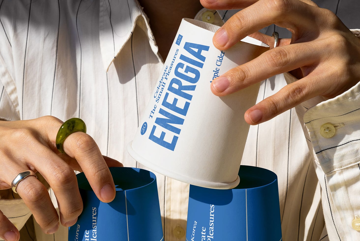 Hands holding white mug with blue ENERGIA cup below it, striped shirt visible