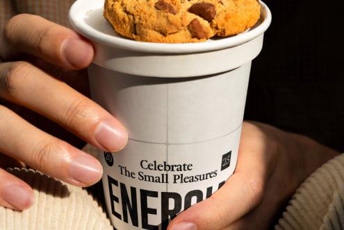 Close up of hand holding white cup with ENERGIA text, brown cookie visible on top