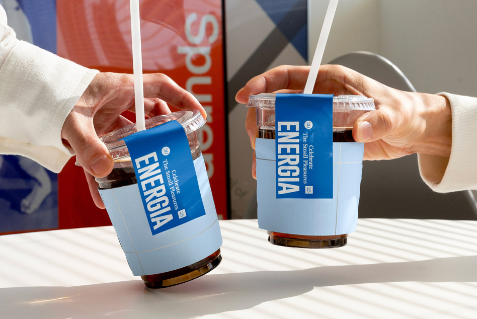 Two people holding clear plastic cups with blue Energia sleeve labels, white straws