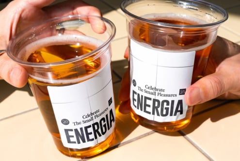 Two hands each holding clear cup with white label showing golden brown drink