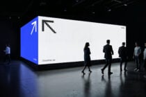 Large exhibition display with blue side panel, white surface, pixel cursor icon, people walking in front