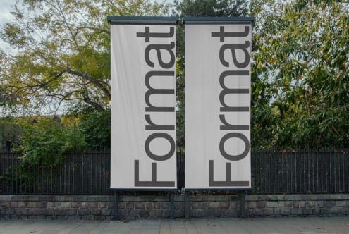 Two white vertical banners on metal frame by tree wall, gray text rotated