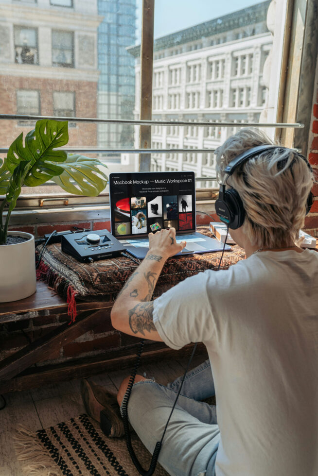 Person with headphones working at desk with MacBook mockup showing design portfolio grid layout near city window