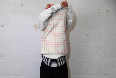 Person wearing white long sleeve with orange grid pattern tank top pulled over head