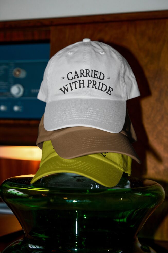 White baseball cap with Carried With Pride text stacked on blue background