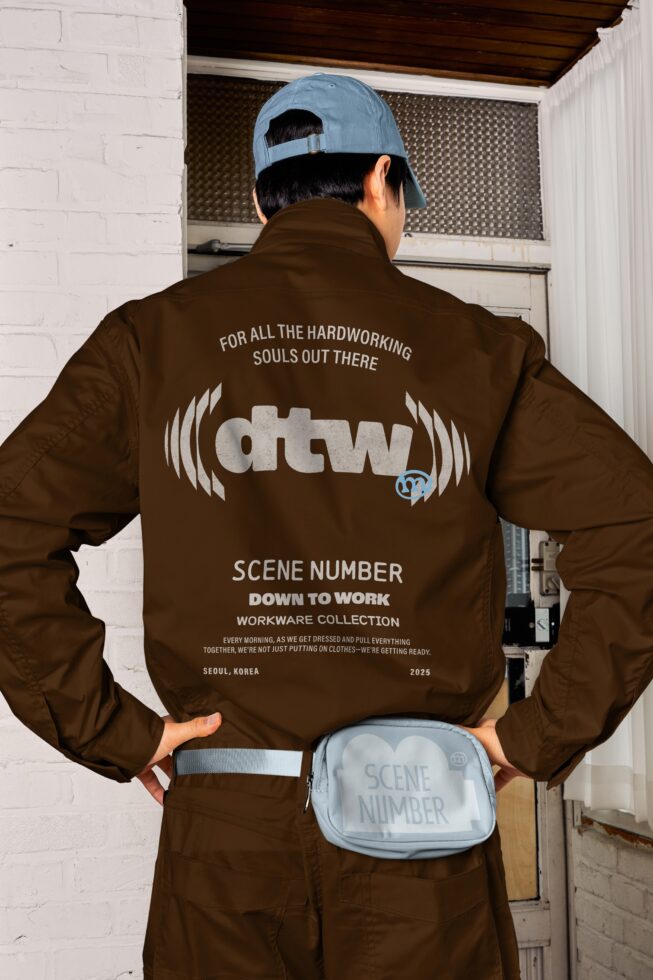 Person wearing brown jacket with DTW logo print, light blue cap, waist bag from Scene Number Down to Work workware line