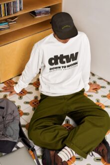 Full body shot wearing white workwear sweatshirt, black cap, olive pants seated