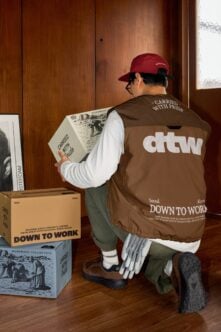 Person carrying boxes wearing maroon cap, brown vest with workwear design visible