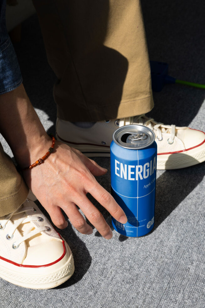 Hand holding blue Energia can next to white sneaker with red stripe on ground