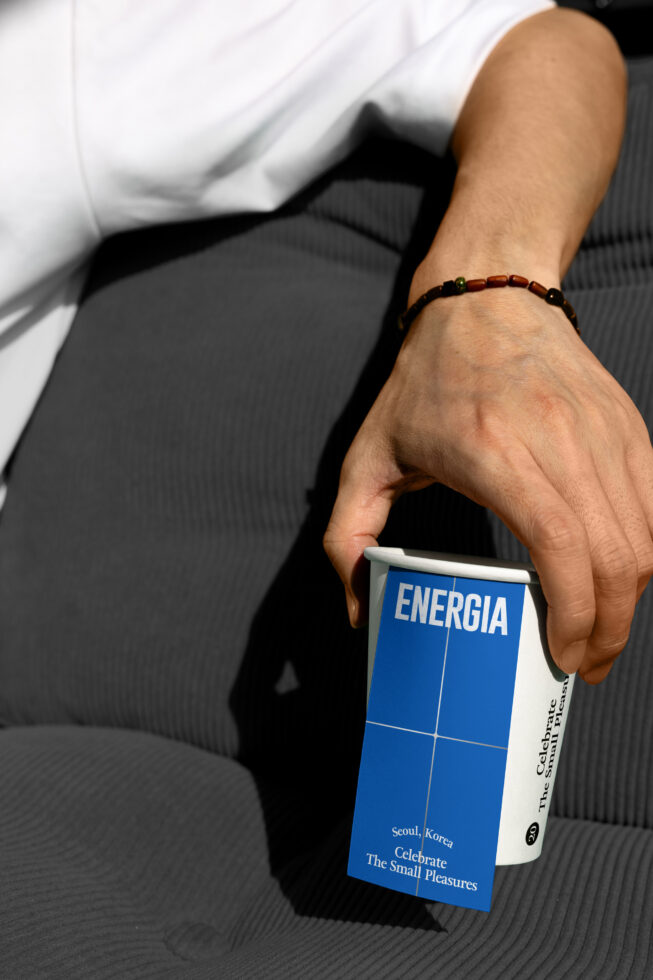 Hand holding paper cup mockup with blue Energia label against dark chair backdrop