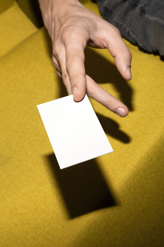 Fingers holding white business card against yellow background with shadow