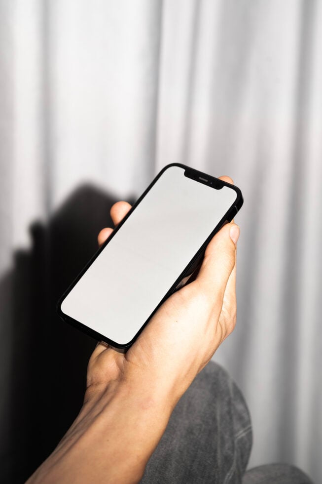 Black smartphone held in hand with blank white screen against white curtain backdrop