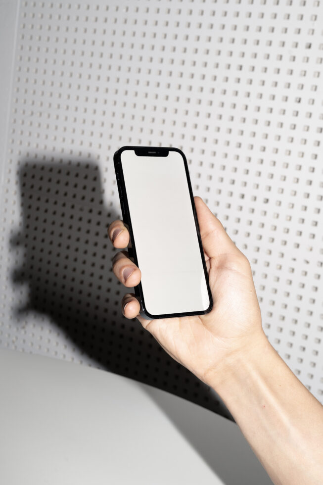 Hand holding black iPhone with blank white screen against perforated wall backdrop