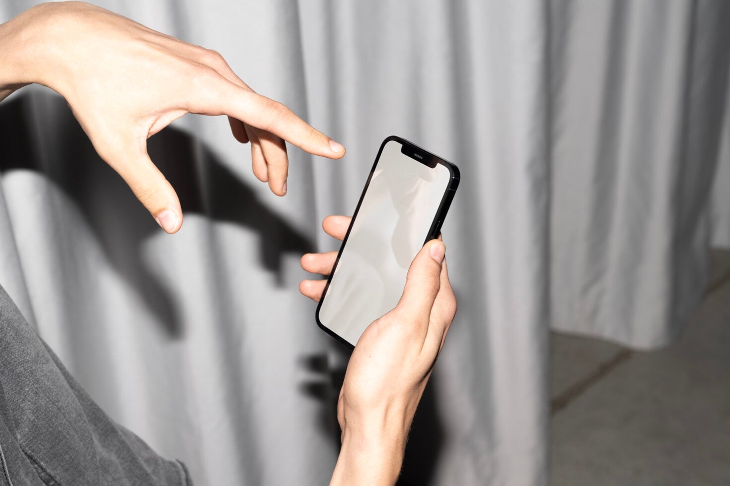 Hands holding black iPhone with blank screen, pointing gesture against white curtain