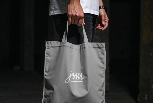 Person holding gray tote bag with small script logo, dark background