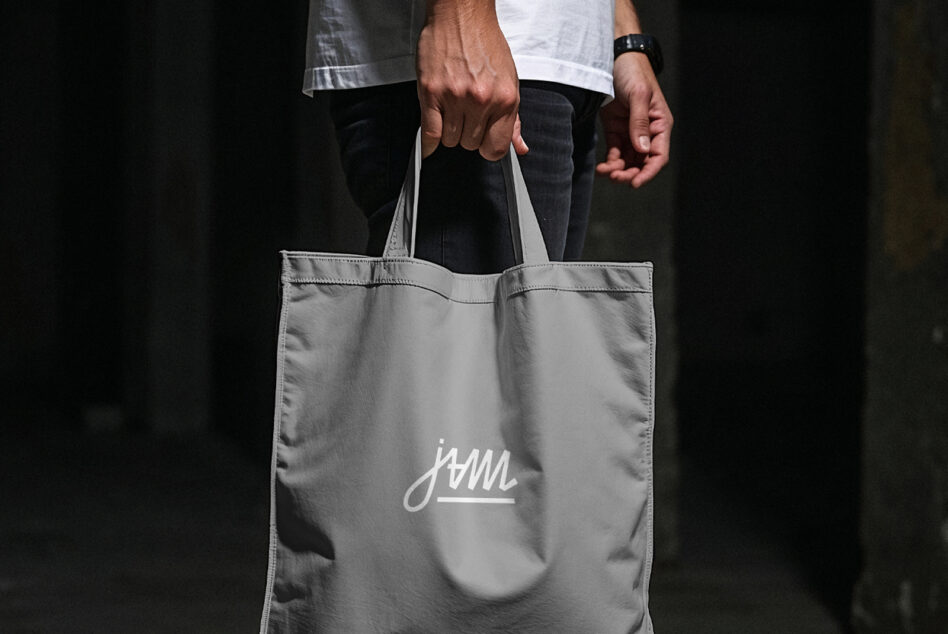 Person holding gray tote bag with small script logo, dark background