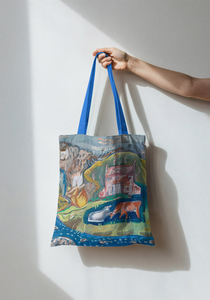 Hand holding blue-handled tote with illustrated rural landscape scene