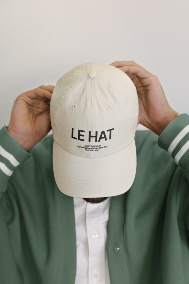 Cream baseball cap with LE HAT text, held up by hands in green jacket