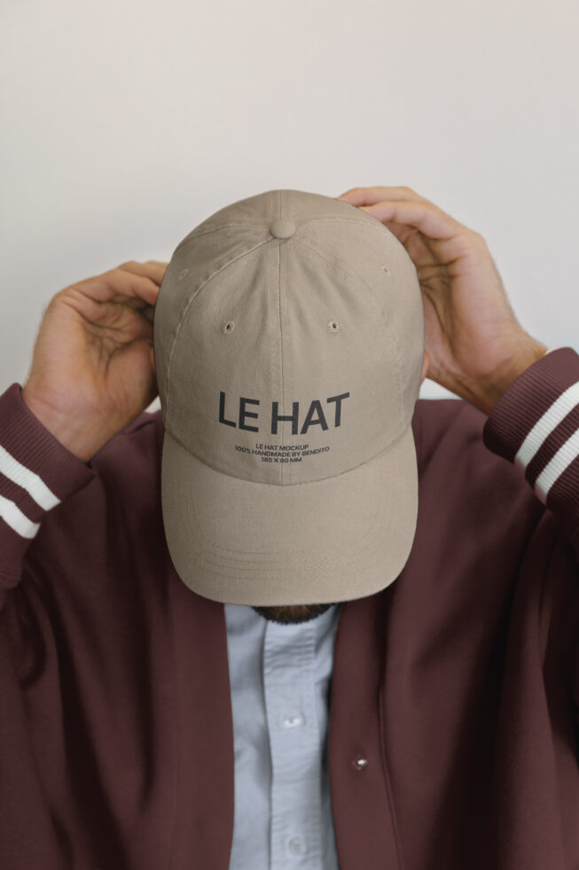 Cream baseball cap with LE HAT text, held up by hands in burgundy jacket