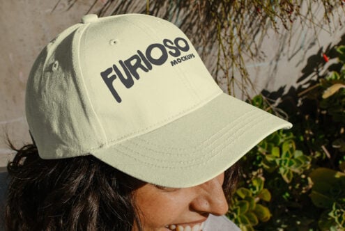Woman wearing cream cap with black Furioso text tilted toward camera near hanging plants