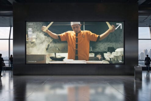 Large indoor screen showing chef in orange shirt tossing noodles with arms raised