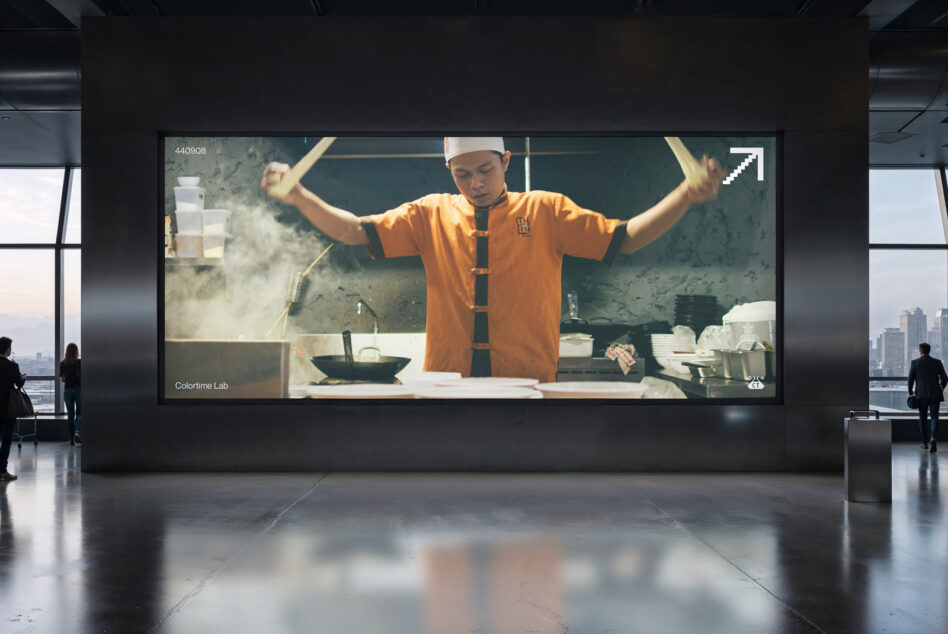 Large indoor screen showing chef in orange shirt tossing noodles with arms raised