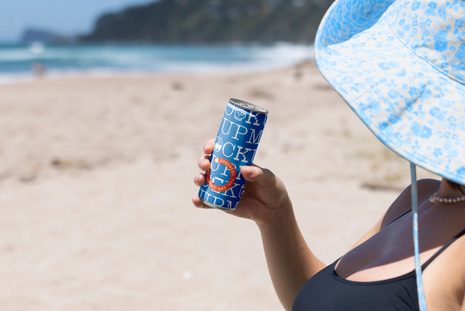 Slim can mockup with blue design held at beach by person under patterned umbrella