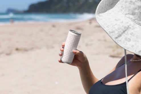 Slim can mockup with grid pattern held at beach by person under white umbrella