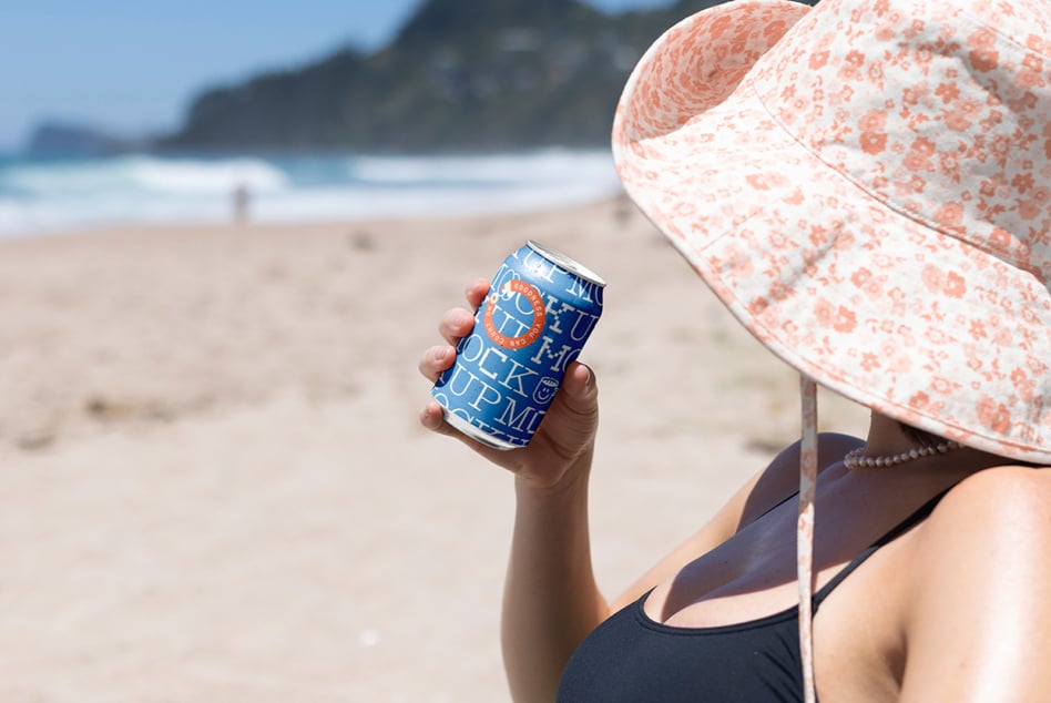 Slim can mockup with blue design held at beach under coral patterned sun hat