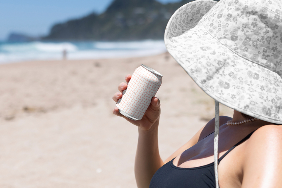 Slim can mockup with grid pattern in hand at beach under patterned sun hat