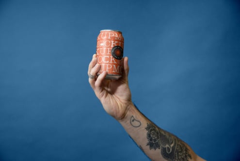 Hand holding orange beer can with repeating text pattern against blue background