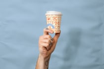 Hand holding branded coffee cup with orange text mockup against sky backdrop