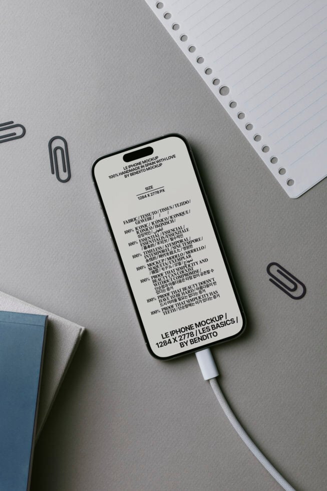 iPhone mockup with white screen showing product specs, USB cable attached, surrounded by office supplies