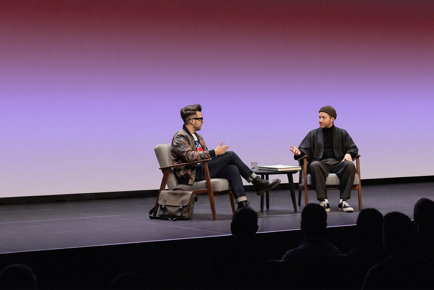 Two men in conversation on stage with purple gradient backdrop, audience silhouettes in foreground