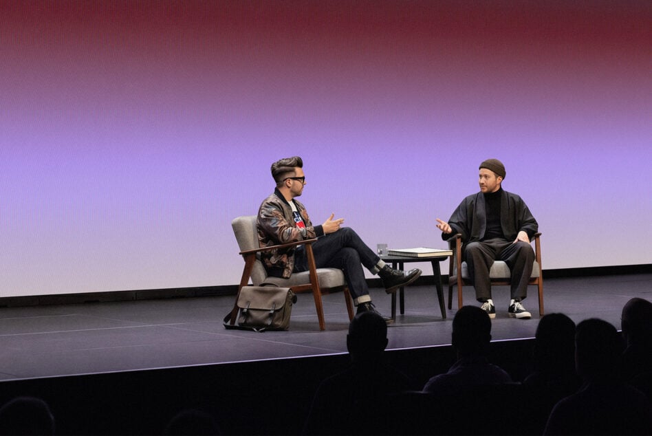 Two men in conversation on stage with purple gradient backdrop, audience silhouettes in foreground