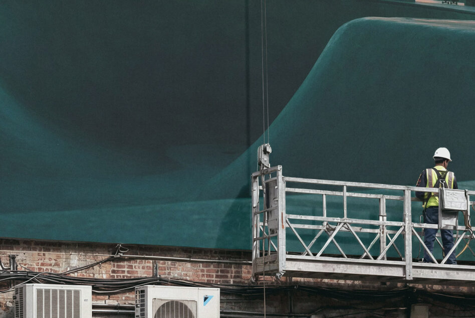Worker on suspended platform next to large teal construction hoarding