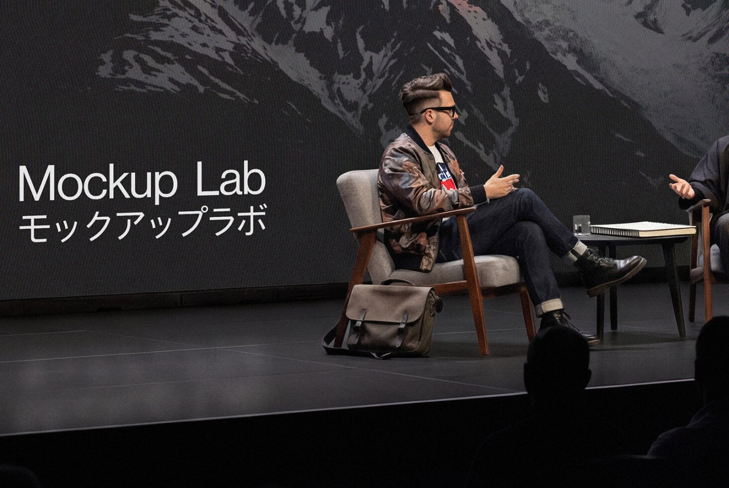 Stage scene with speaker in chair talking, large screen behind showing Mockup Lab text
